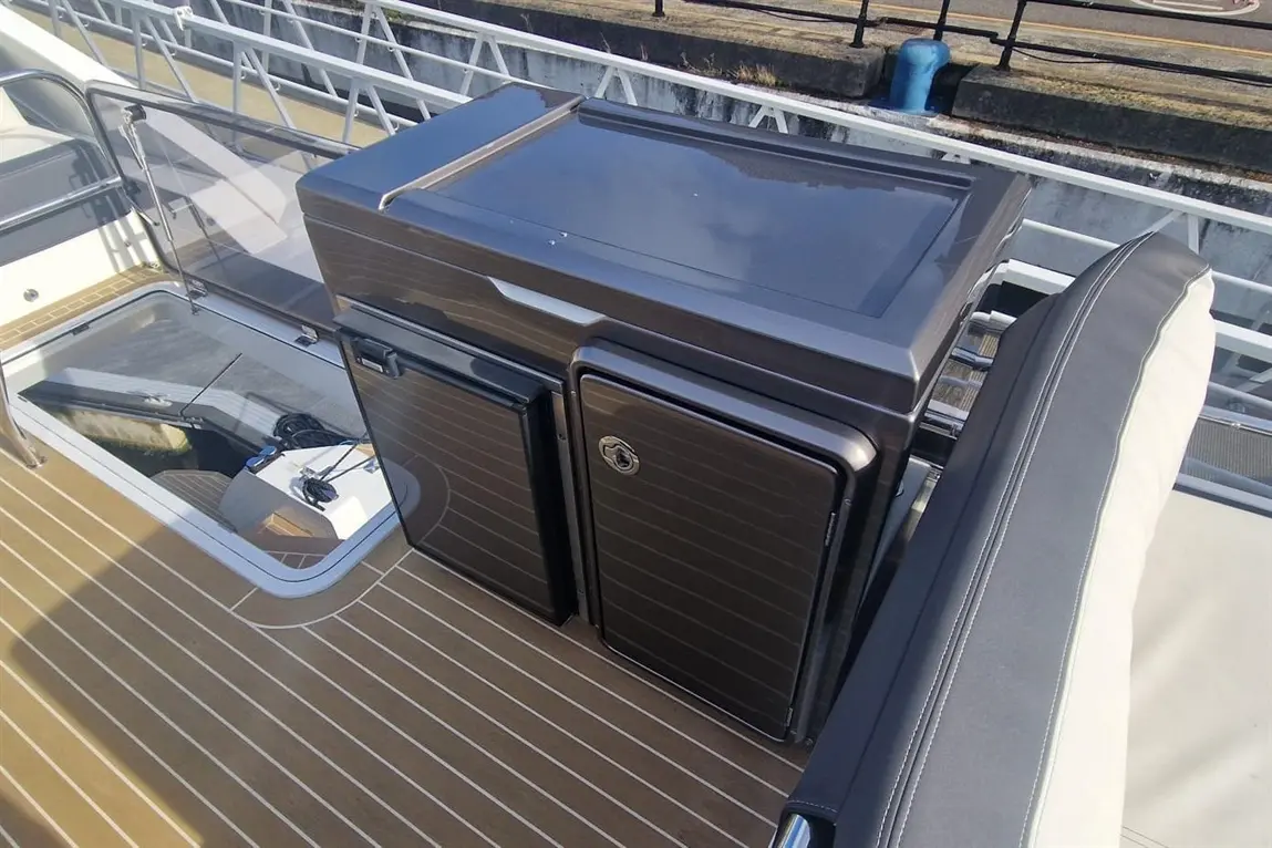 A portable marine fridge sits on a boat deck, alongside a transparent hatch and a cushioned seat with grey upholstery.